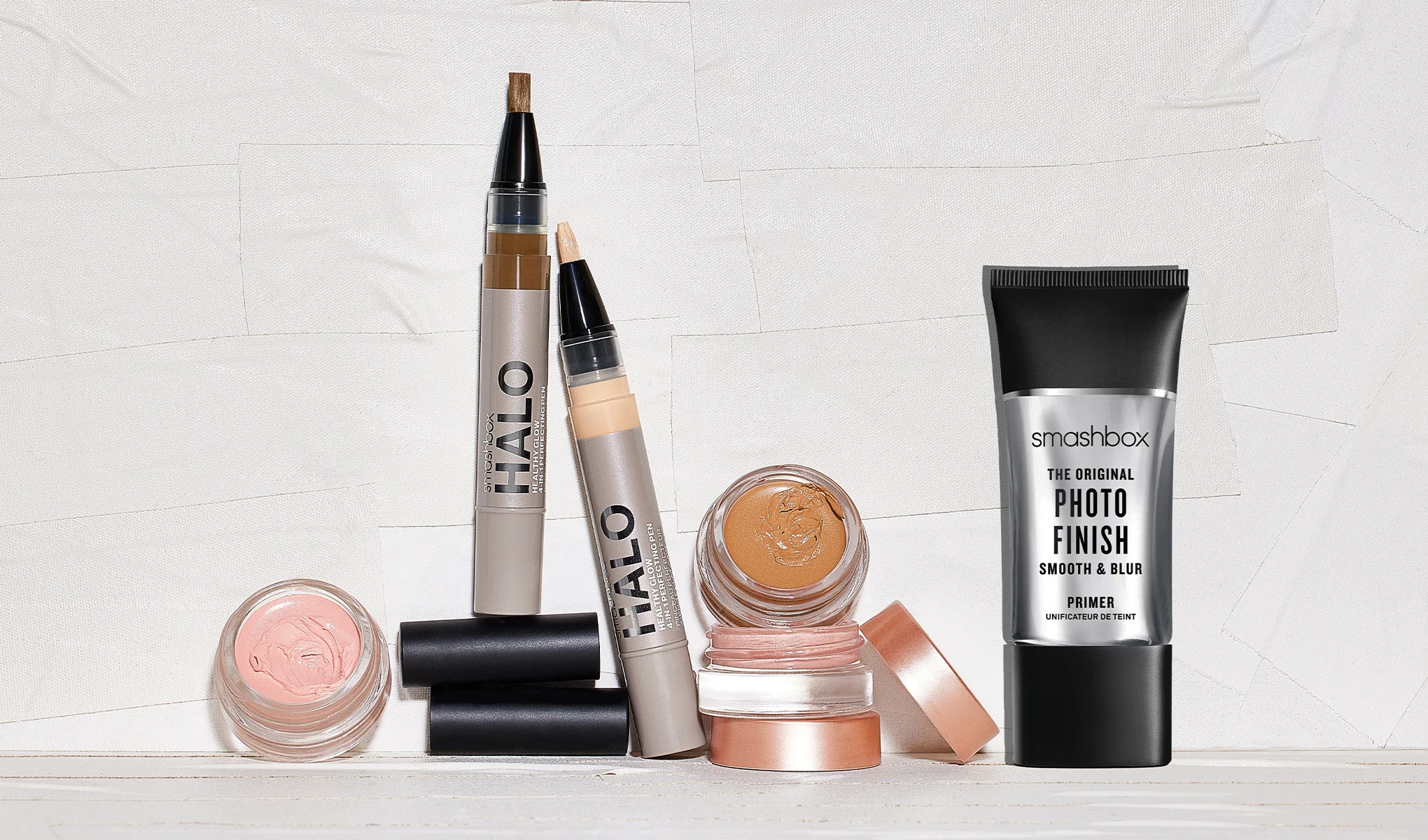 En samling sminkprodukter från Smashbox placerade på en vit yta mot en ljus bakgrund. Produkterna inkluderar en svart tub, en öppen palett, en beige tub, en liten beige tub och en svart penna. Produkterna har förpackningar i svart, beige och rosa.