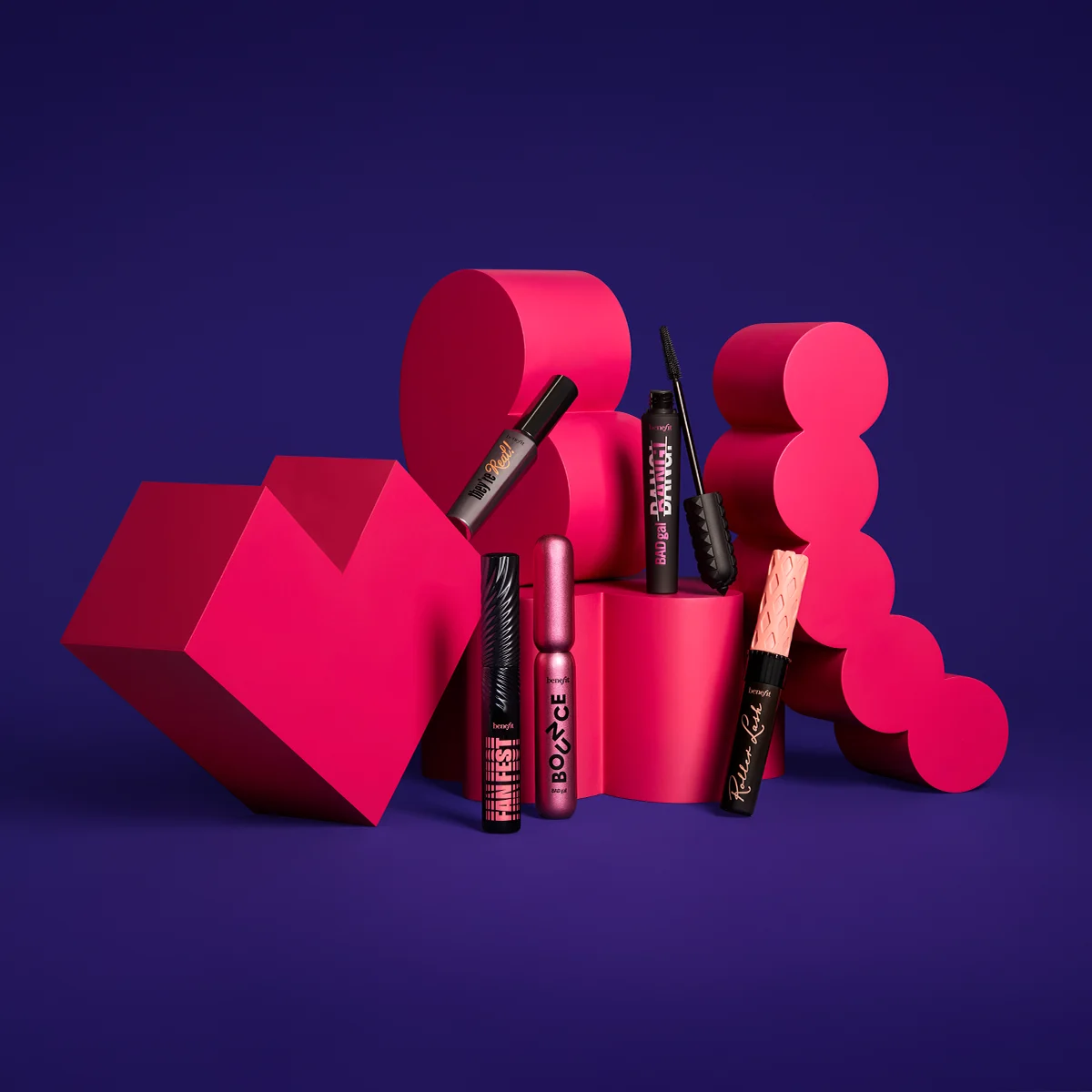Fem olika mascaror från Benefit Cosmetics visas mot en färgstark bakgrund med geometriska former i rosa och lila. Produkterna har en unik och färgstark design.