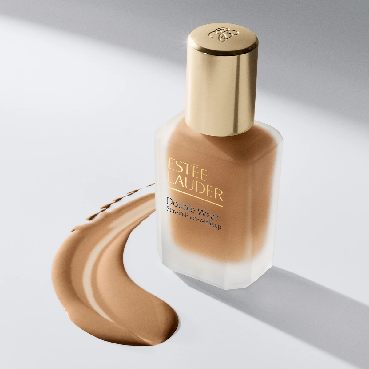 Estée Lauder Double Wear Stay-in-Place Makeup foundation i beige nyans med guldfärgad kork, placerad bredvid ett svep av flytande foundation.