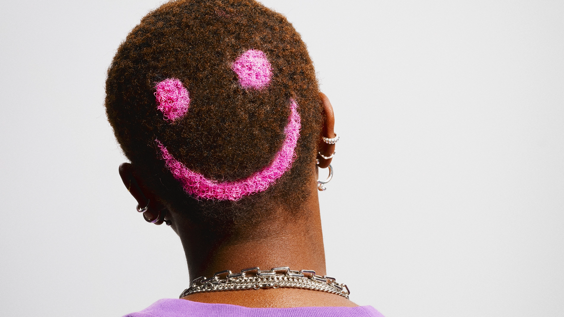 Bakhuvud med kort, lockigt hår där ett rosa smiley-motiv är infärgat i håret. Personen bär lila tröja, flera örhängen och ett silverfärgat halsband.