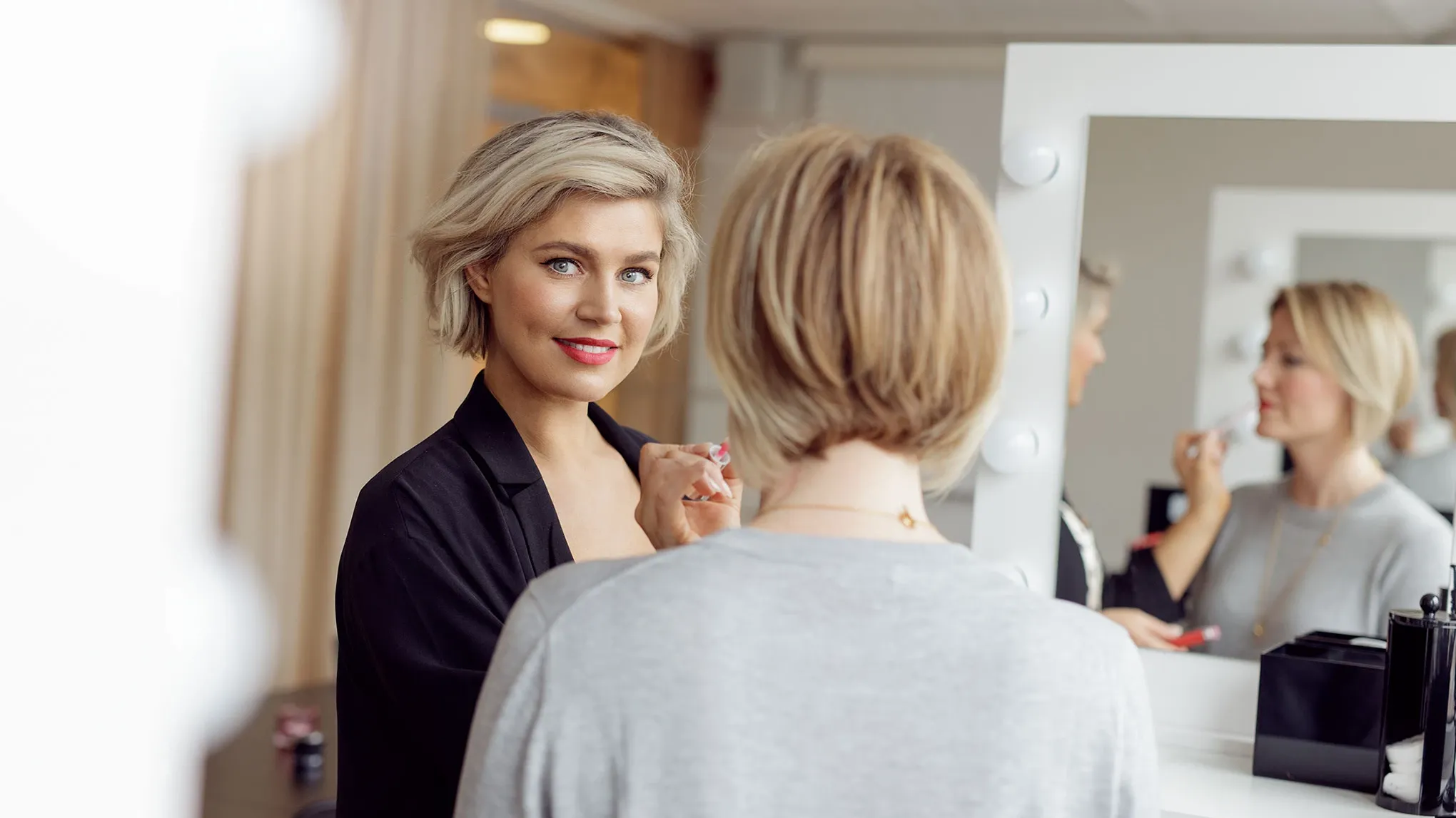 Blond kvinna i fokus applicerar läppstift på en annan kvinna med ryggen mot kameran, i ett rum med beiga gardiner, vita väggar och en stor spegel.