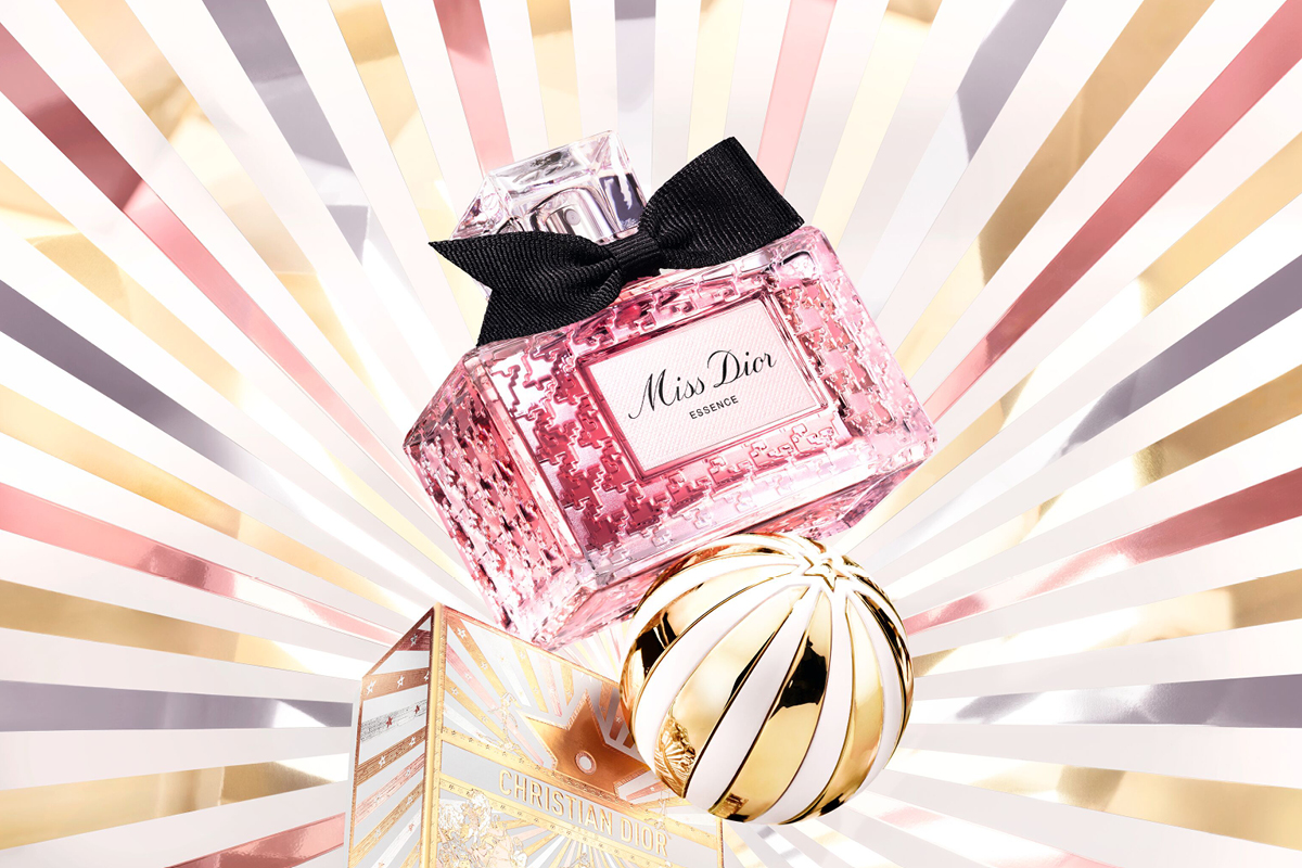 Vaaleanpunainen Miss Dior Essence -pullo, taustalla säteilevät linjat kultaisena, vaaleanpunaisena ja valkoisena, sekä Christian Dior-lahjapakkaus ja koristeellinen pallo.