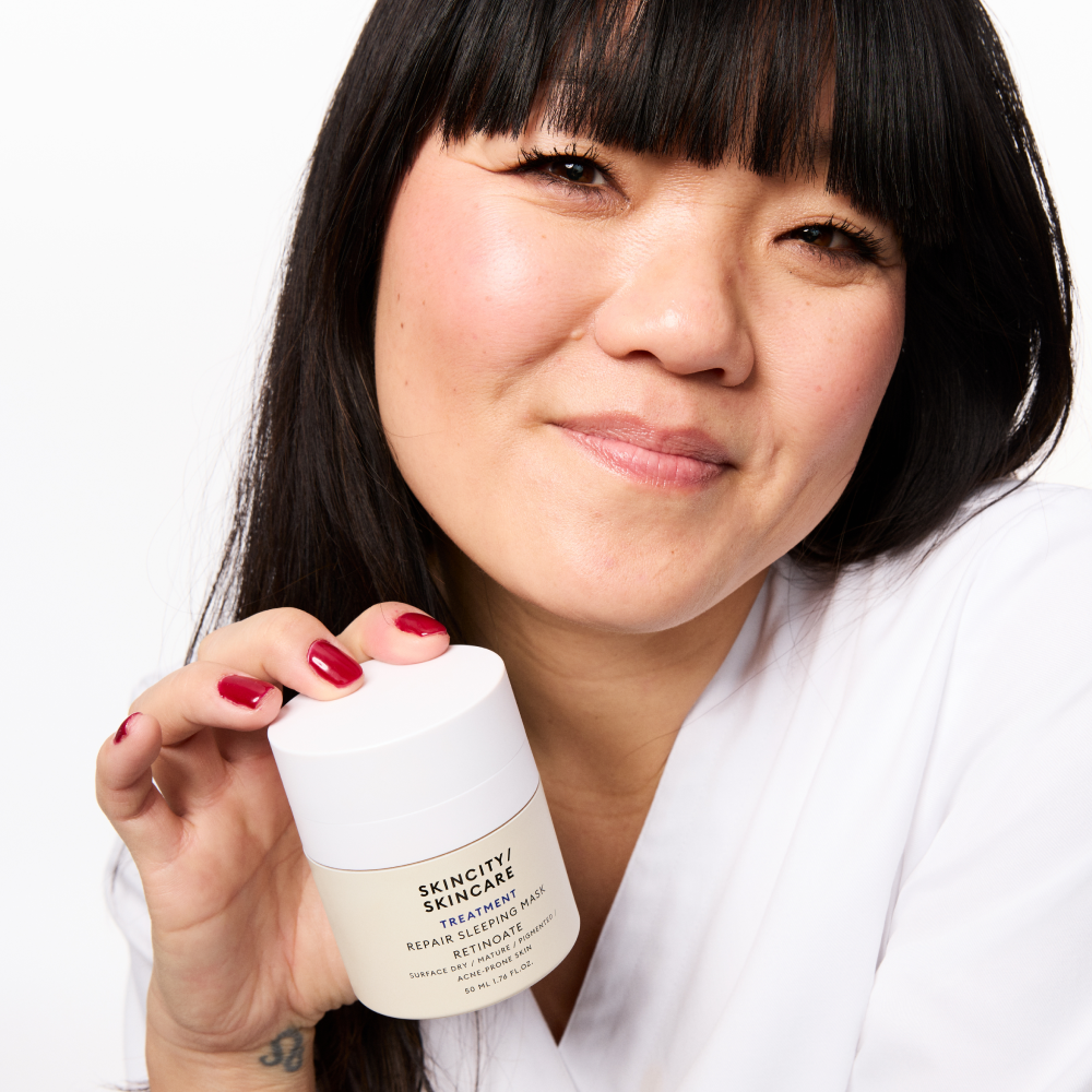 Henkilö, jolla on tummat hiukset ja punaiset kynnet, pitää kädessään Skincity Skincare Repair Sleeping Mask -purkkia. Purkki on valkoinen ja siinä on musta teksti, ja henkilöllä on yllään valkoinen paita.