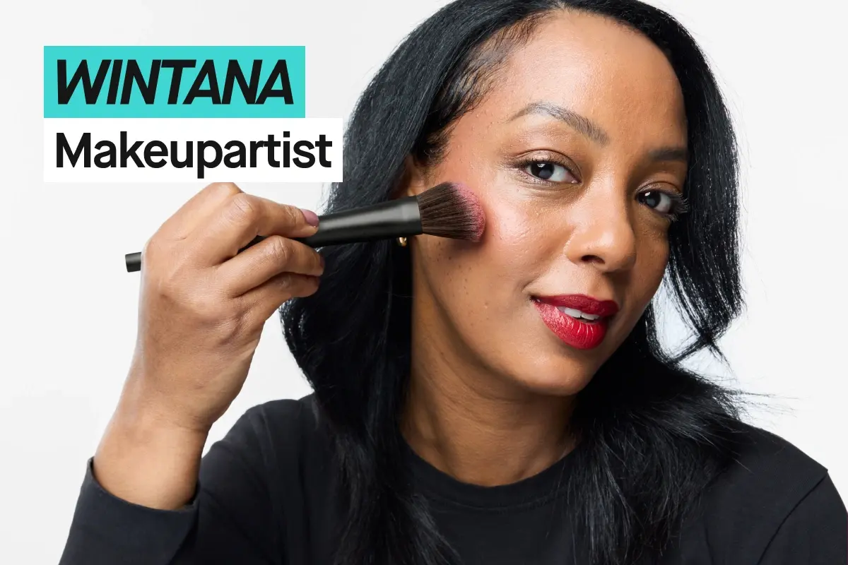 Person med svart hår påfører blush med sminkekost på kinnet mot lys grå bakgrunn og har på seg en svart KICKS-t-skjorte. Turkis og hvit tekst viser “WINTANA” og “Makeupartist”.