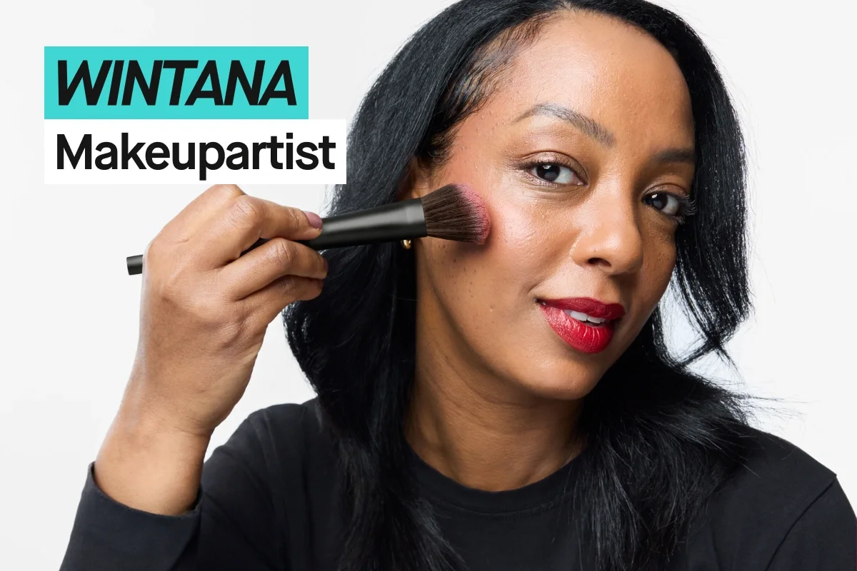 Person med svart hår påfører blush med sminkekost på kinnet mot lys grå bakgrunn og har på seg en svart KICKS-t-skjorte. Turkis og hvit tekst viser “WINTANA” og “Makeupartist”.