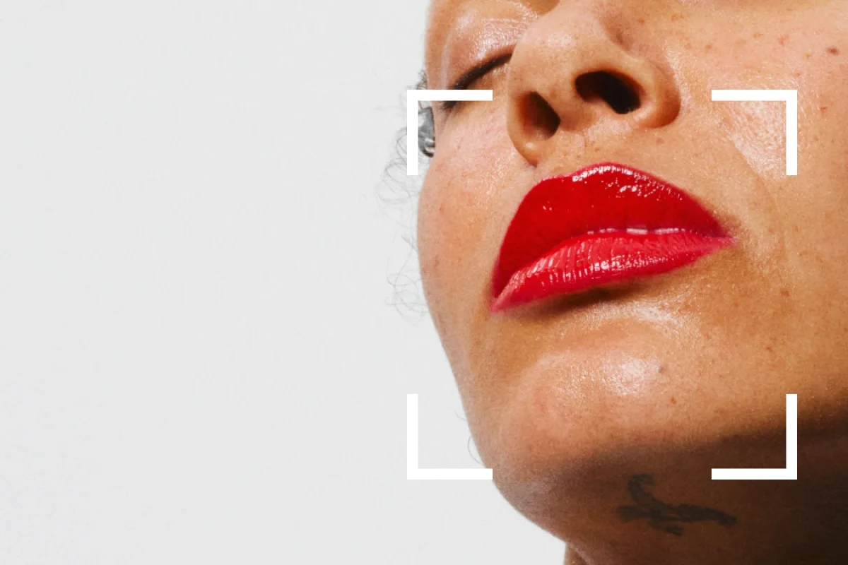Nærbilde av en persons lepper med glansfull rød leppestift, med fokusram-grafikk rundt ansiktet mot en lys bakgrunn. Hudens tekstur og glød er synlig i denne makeup-looken med leppestift.