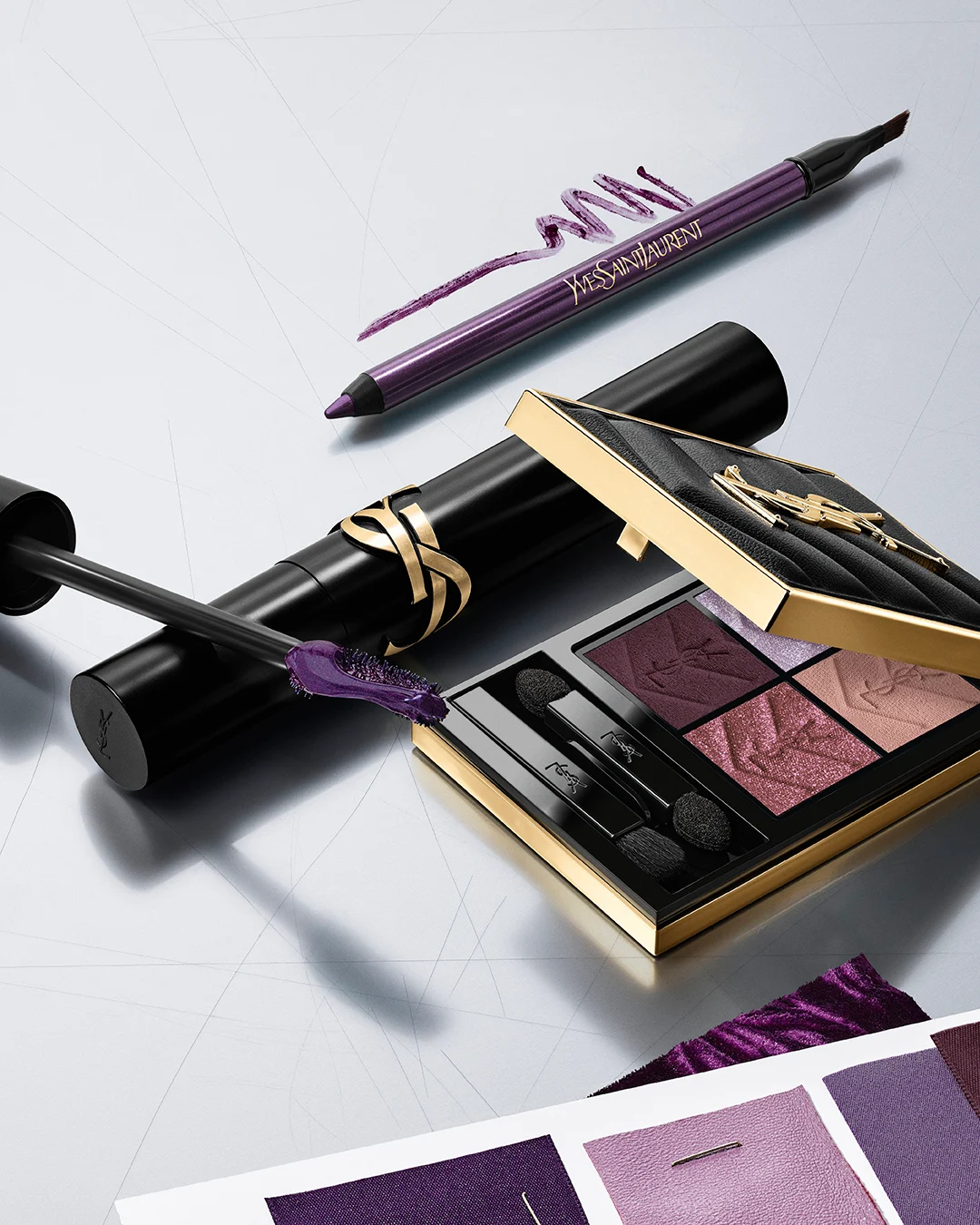Nærbilde av en person med skimrende lilla øyesminke, rosa highlighter og glansfulle lepper. Personen holder et svart Yves Saint Laurent-produkt med gullfarget YSL-logo og bærer gullfargede øredobber mot lys bakgrunn.