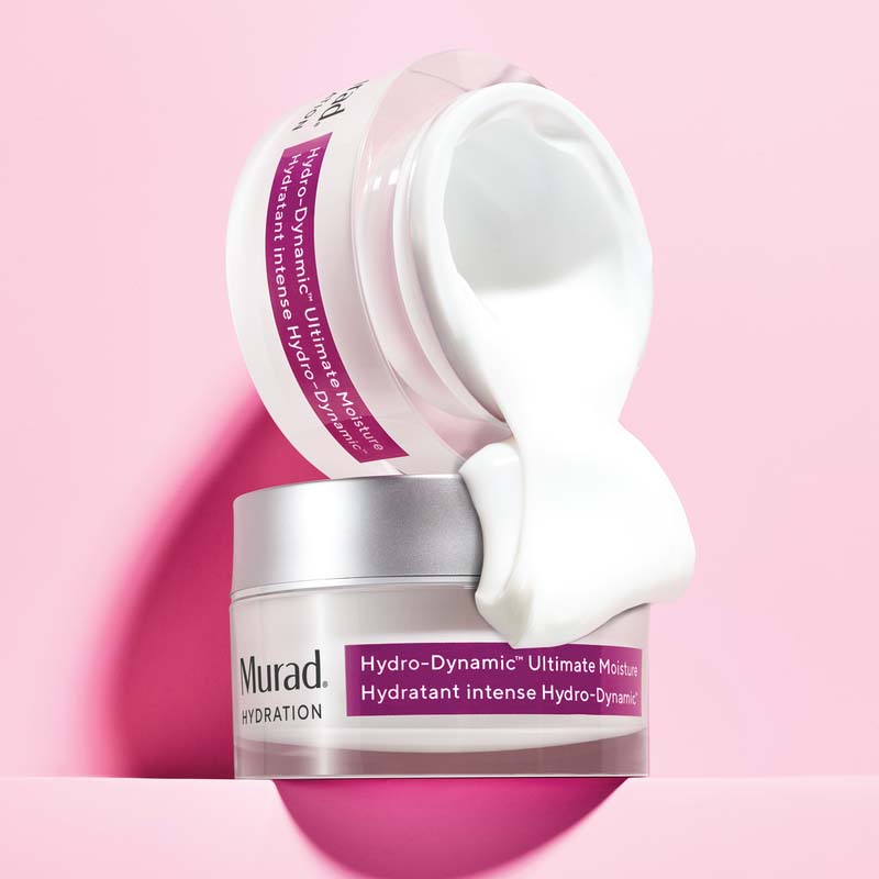 Två burkar nattkräm från Murad. Den ena brukar är öppen med vit kräm är placerad ovanpå en annan burk mot en rosa bakgrund. Den övre burken har en lila etikett som visar produktnamnet och krämens innehåll rinner ner över den nedre burken, vilket framhäver dess rika och återfuktande konsistens.