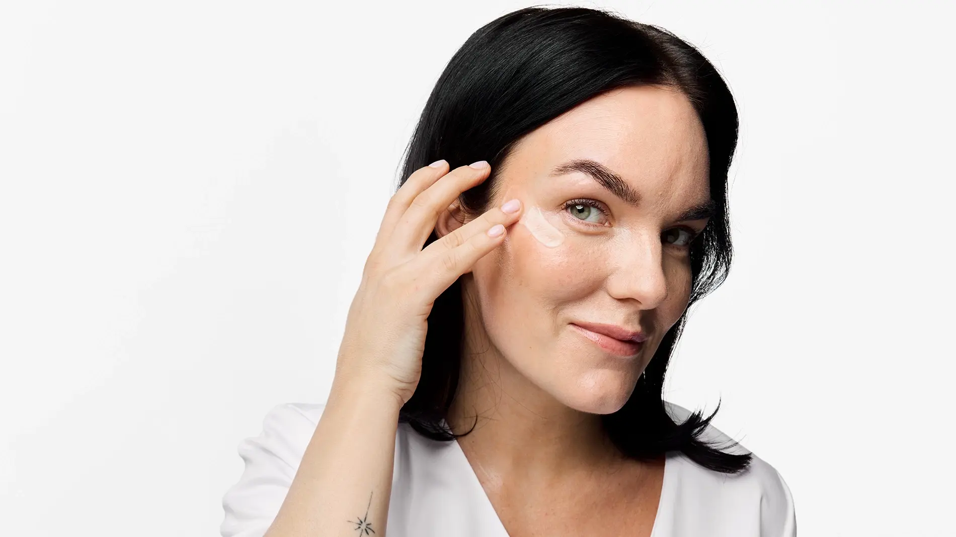 Person påfører øyekrem eller hudpleiekrem under øyet med fingertuppene. Nærbilde med naturlig makeup, mørkt hår og hvit topp mot lys bakgrunn.