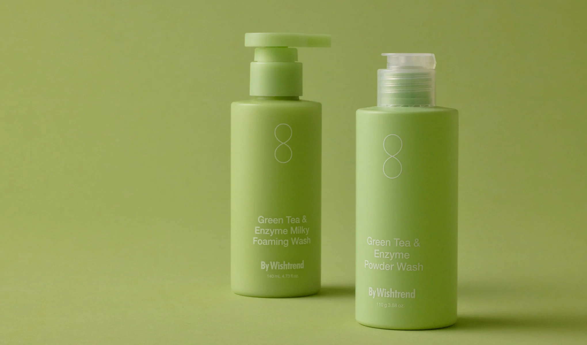 To grønne flasker med By Wishtrends Green Tea & Enzyme Milky Foaming Wash og Green Tea & Enzyme Powder Wash, plassert mot en grønn bakgrunn. Flaskene har hvite etiketter og tekst.
