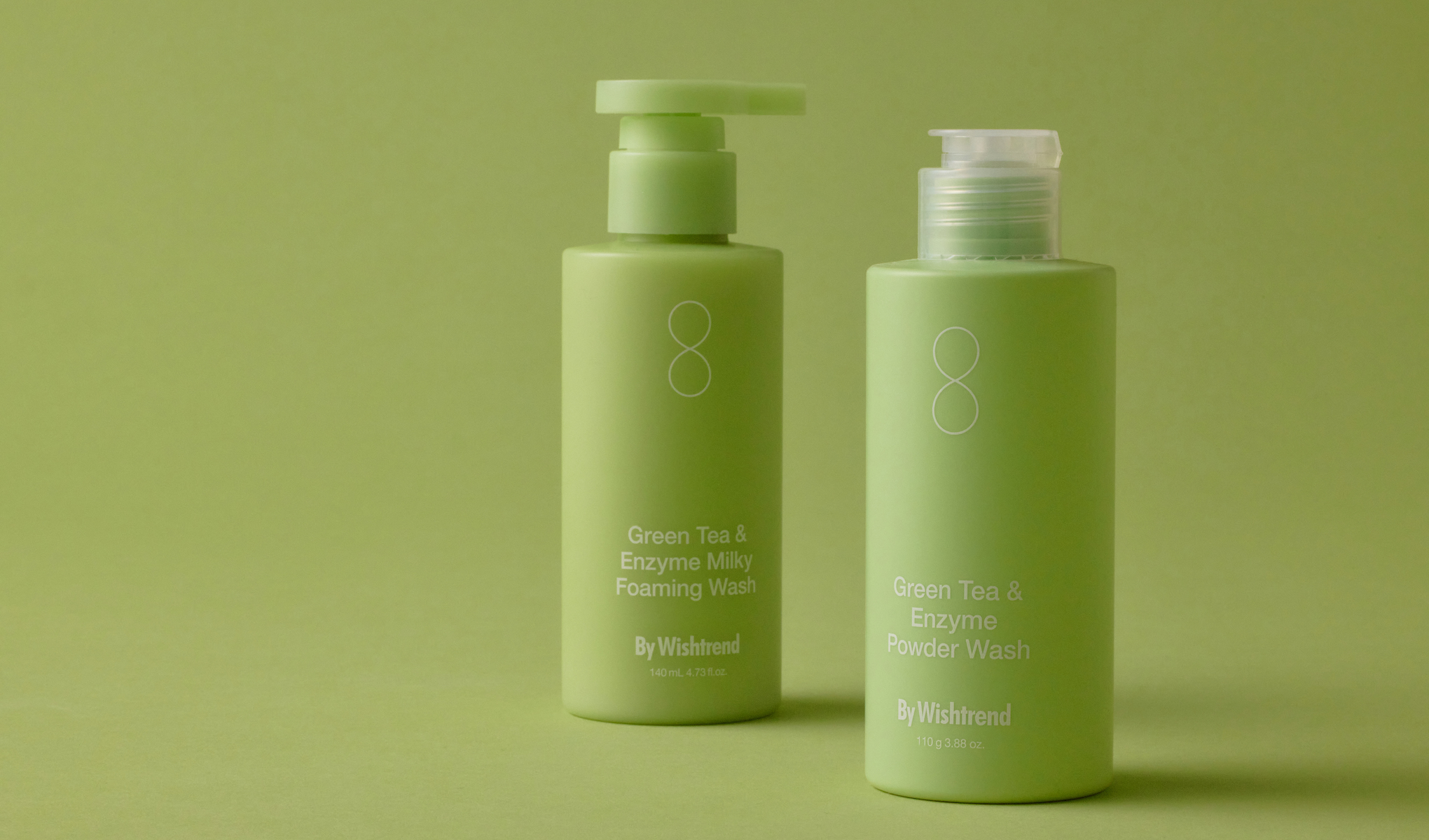 Kaksi vihreää pulloa By Wishtrendsin Green Tea & Enzyme Milky Foaming Wash -vaahtopesua ja Green Tea & Enzyme Powder Wash -jauhepesuaineita vihreää taustaa vasten. Pulloissa on valkoiset etiketit ja teksti.