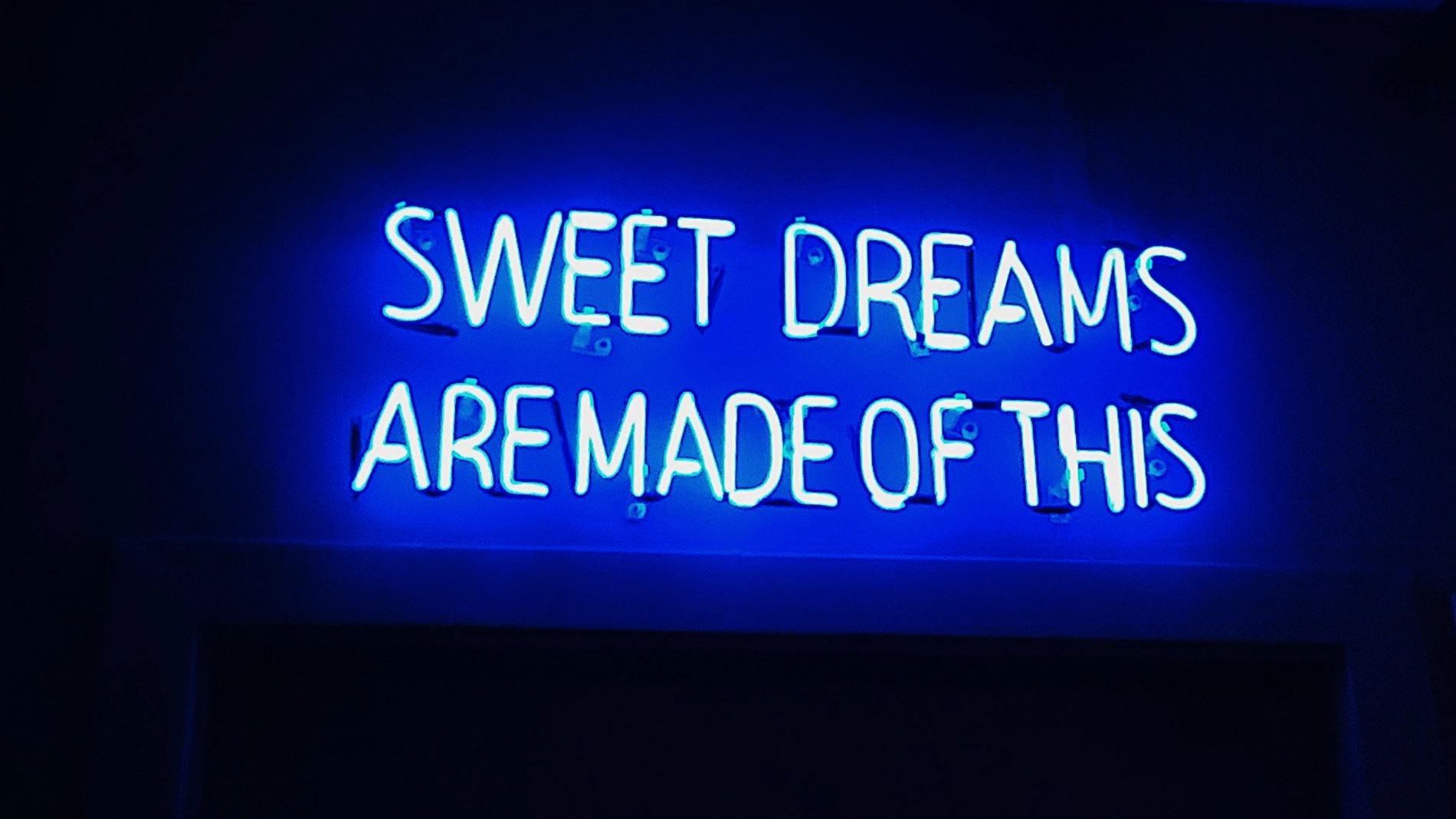 Sininen neonkyltti tummalla seinällä, jossa näkyy valaistu teksti ”SWEET DREAMS ARE MADE OF THIS”. Kyltti luo sinistä hohdetta ympärilleen ja on kuvan keskeinen elementti.