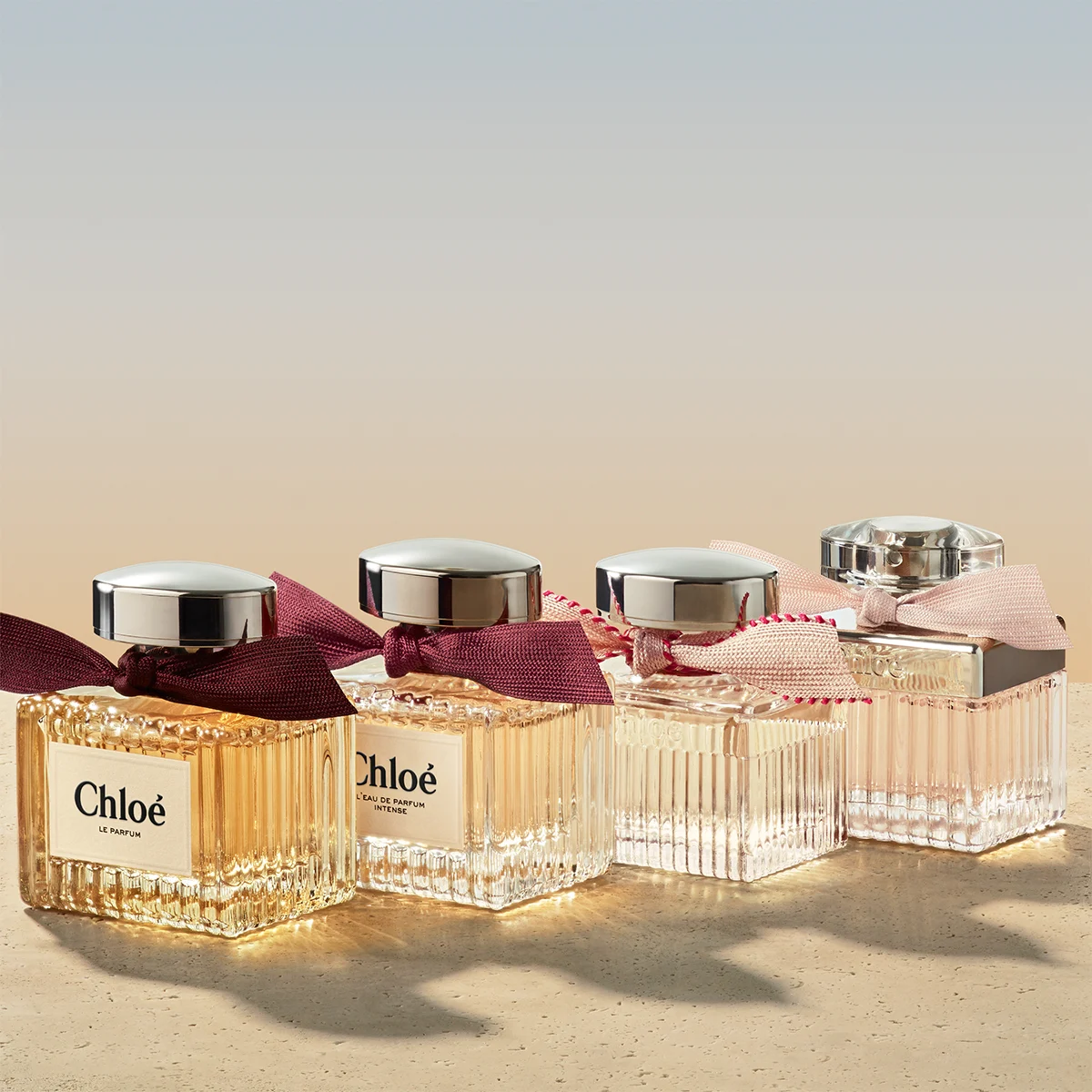 Tre parfymflaskor från Chloé. Ena flaskan är dekorerad med en mörkröd rosett och har en silverfärgad kork. De andra två är dekorerade med ljusrosa rosett. De är placerad på en ljus yta med mjuk belysning som skapar reflektioner. Bakgrunden är ljus och neutral.