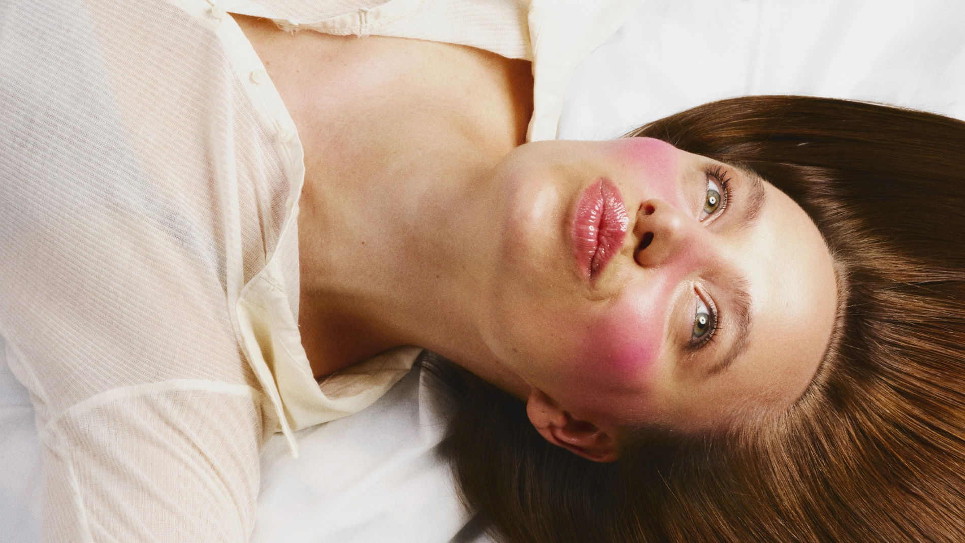 En person ligger på ryggen på hvitt sengetøy med langt, brunt hår, rosa kinn og en lys skjorte.