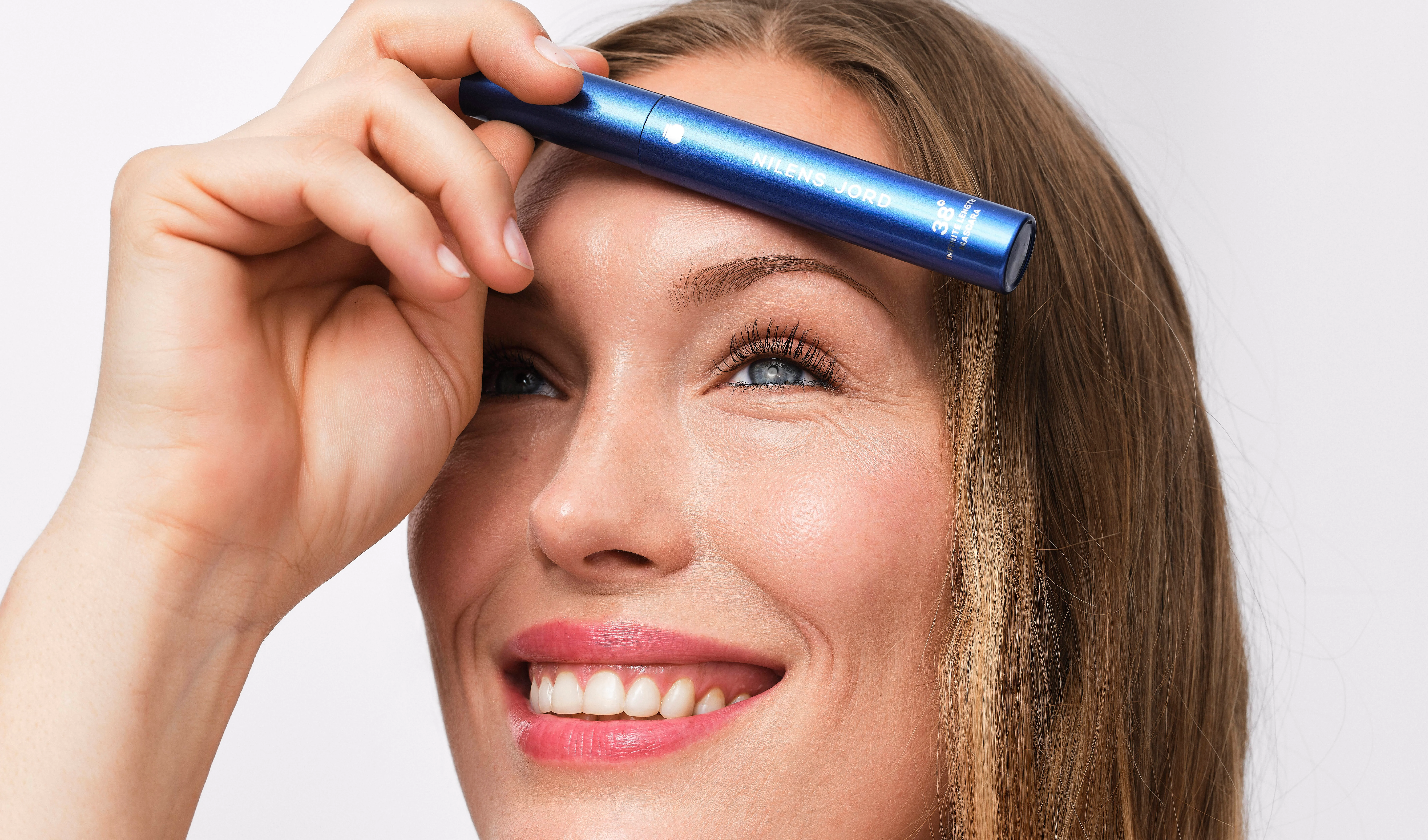 Person håller en blå mascara från Nilens Jord framför ansiktet och visar långa separerade fransar med naturlig ögonmakeup. På förpackningen syns texten NILENS JORD och “38° Infinite Lash Mascara”.