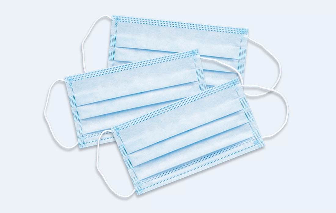 Tre blå engangsmunnbind med hvite ørestrikker liggende på en lys bakgrunn.