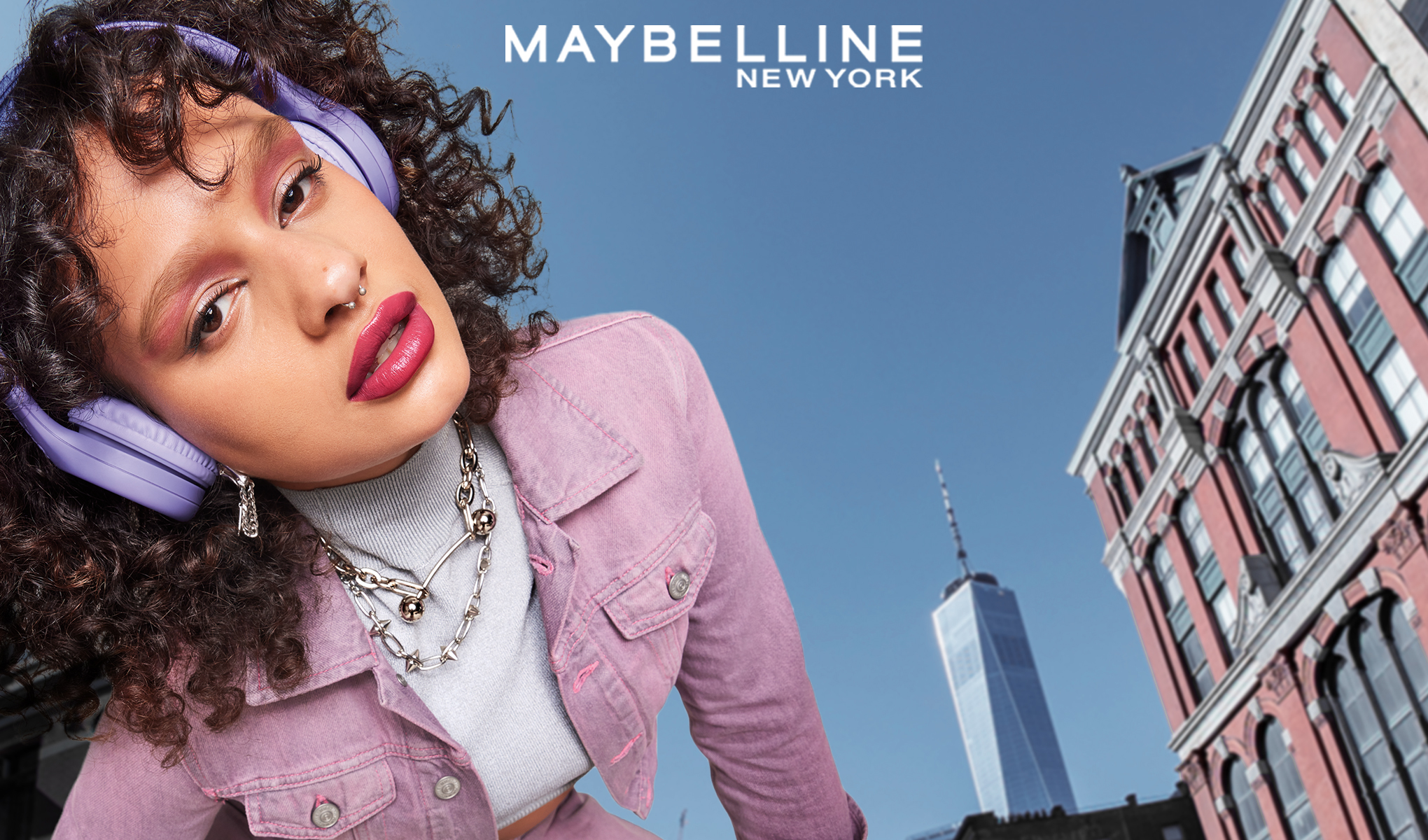 En kvinna med lockigt hår, som bär lila hörlurar och en rosa jacka. Ovanför henne står Maybelline New York i vitt. I bakgrunden syns en blå himmel och en byggnad.