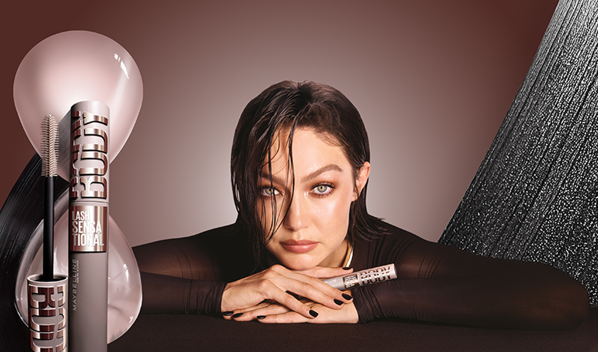 Maybelline Lash Sensational Firework -ripsiväri esillä, malli nojaa käsiinsä ruskealla taustalla.