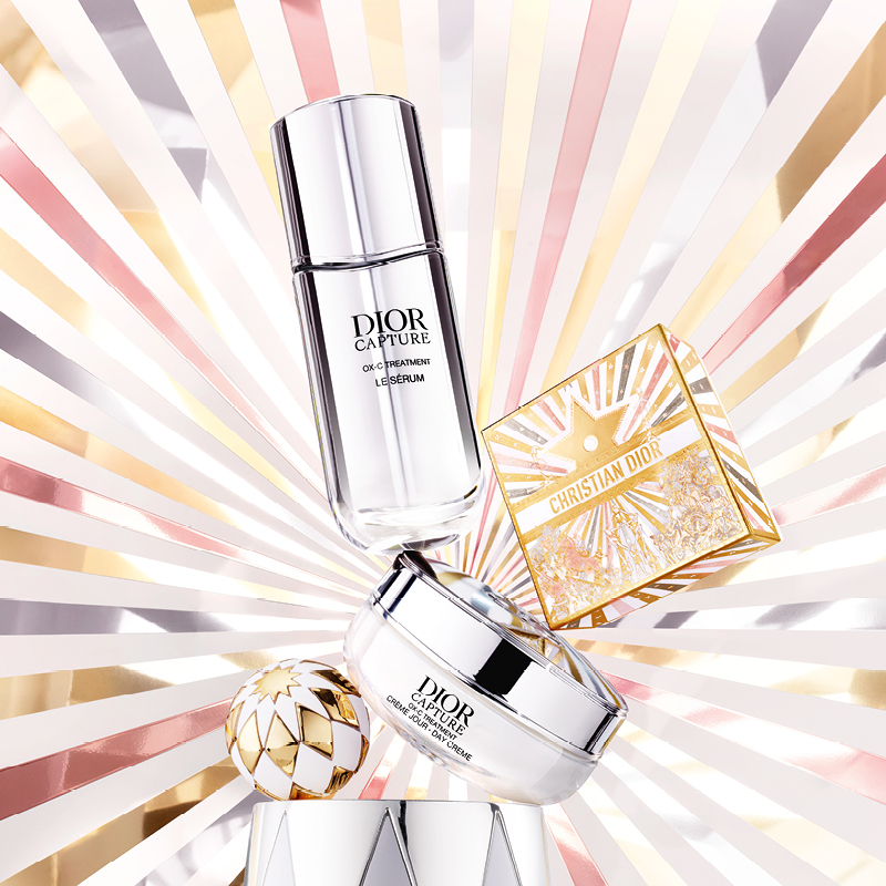 Dior Capture-serumflaska och krämbehållare, med en bakgrund av strålande linjer i guld, rosa och vitt, samt en Christian Dior-presentförpackning och en dekorativ kula.