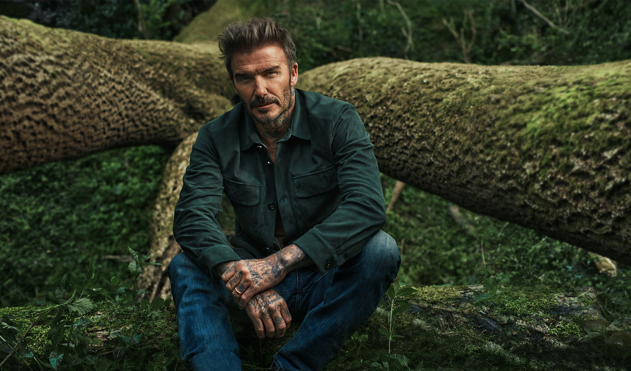 Mies istuu sammaleisen kaatuneen puun päällä metsässä ja katsoo suoraan kameraan.