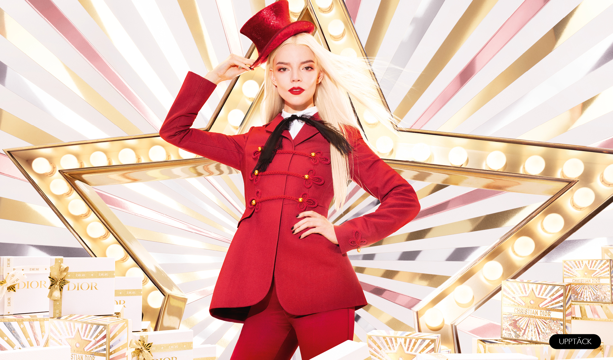 En person klädd i röd kavaj och byxor, med en röd hatt i handen. I bakgrunden finns en stor upplyst stjärna och Dior-presentförpackningar.