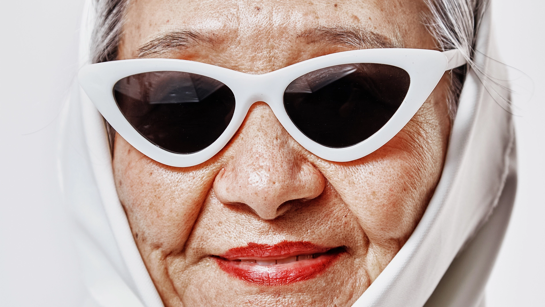 Nærbilde av en person med hvite solbriller og rød leppestift, med fokus på hud og lepper mot en lys, minimalistisk bakgrunn.