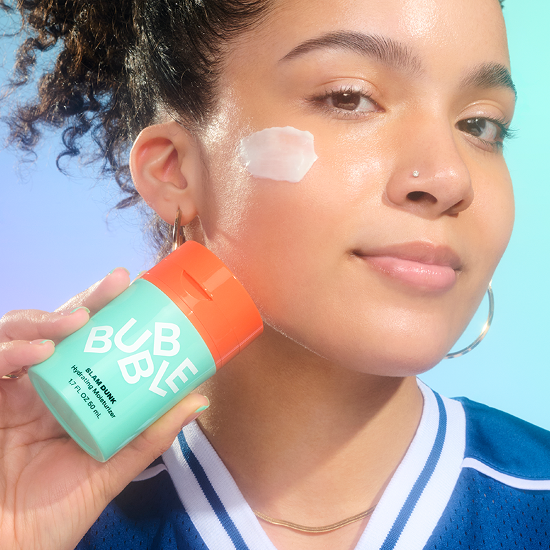 En person holder Bubble Slam Dunk fuktighetskrem i en mintgrønn flaske med oransje kork. Bakgrunnen er blågrønn.