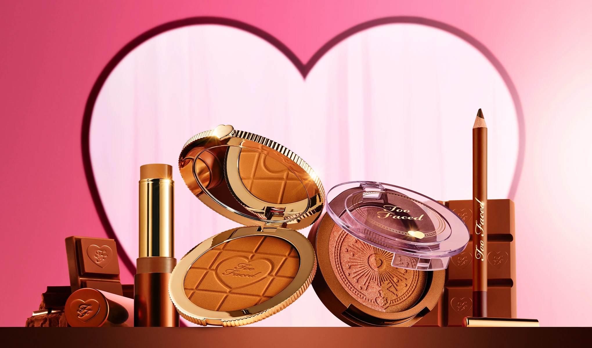 Too Faced -meikkituotteita sydämenmuotoisen taustan edessä, mukana bronzer, puuteri ja meikkikynä.