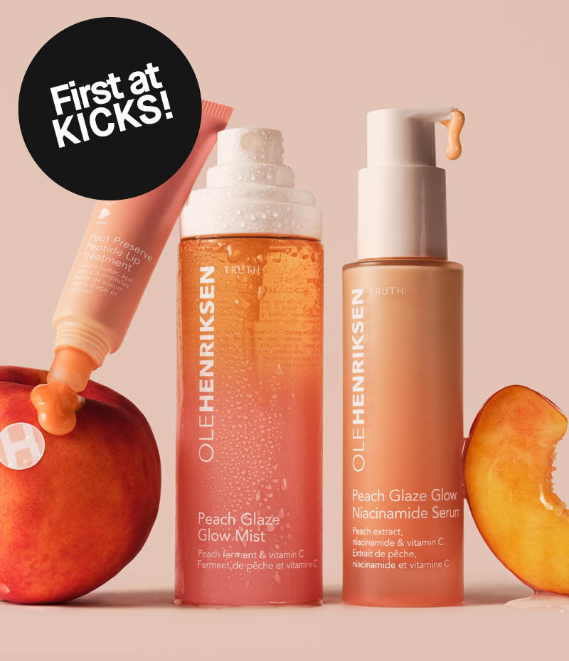 Kolme ihonhoitotuotetta Ole Henriksenin Peach Glaze -sarjasta – huulihoito, suihke ja seerumi – persikanvärisellä taustalla, tuoreiden persikoiden kera. Musta tarra, jossa lukee First at Kicks!
