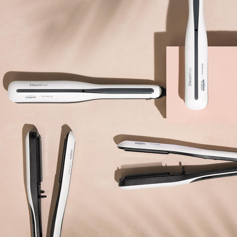 Viisi valkoista L'Oréal Professionnel SteamPod -suoristuslaitetta on levitetty beigeen pintaan. Osa suoristusraudoista on avoinna ja osa kiinni. Taustalla on varjostettu vaikutus.