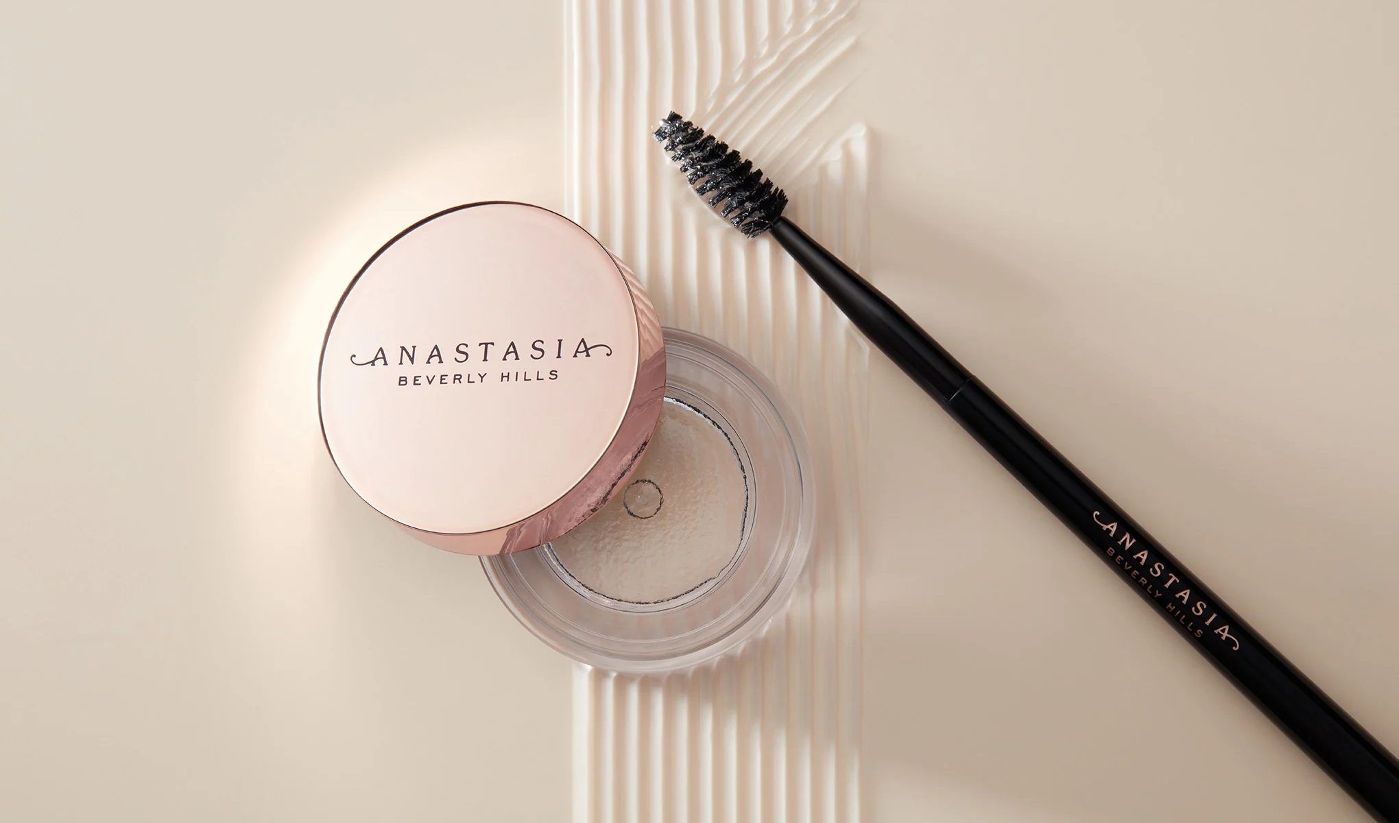 Ögonbrynsprodukt från märket Anastasia Beverly Hills. Locket har texten "Anastasia Beverly Hills" tryckt på sig. Borsten ligger bredvid burken och har samma text tryckt på skaftet. Bakgrunden är ljus och krämfärgad, med en texturerad yta under produkterna som skapar ett elegant och stilrent intryck.