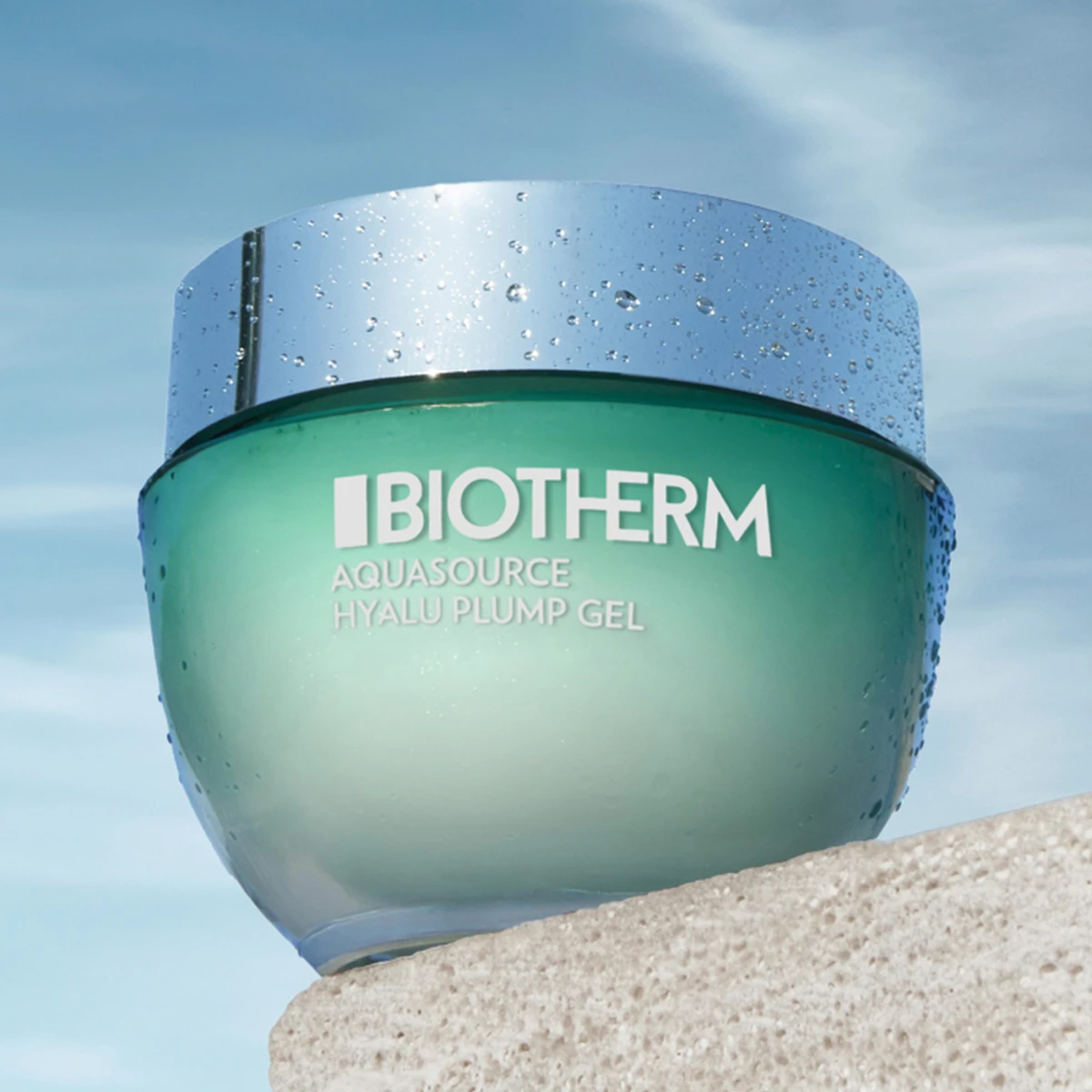 Aquasource Hyalu Plump -geeli Biothermiltä. Geeli on vihreässä lasipurkissa, jossa on hopeinen kansi ja jonka etupuolella on teksti ”Biotherm Aquasource Hyalu Plump Gel”. Purkki seisoo beigen värisellä alustalla sinistä taivasta vasten, jossa on joitakin valkoisia pilviä.
