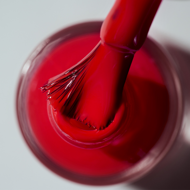 En nagellacksborste doppad i rött nagellack. Borsten är i närbild och nagellacksflaskan är delvis synlig. Bakgrunden är ljusgrå.