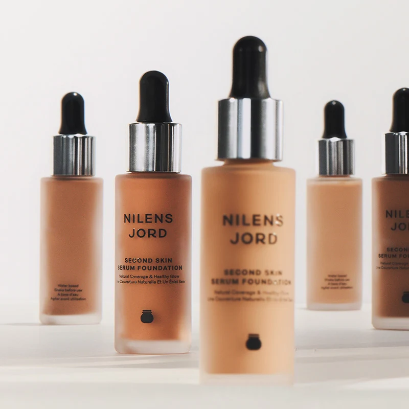 Nilens Jord Second Skin Serum Foundation. Flera flaskor med beige foundation och svart pipett, samt silverfärgad hatt. Flaskorna har svart text.