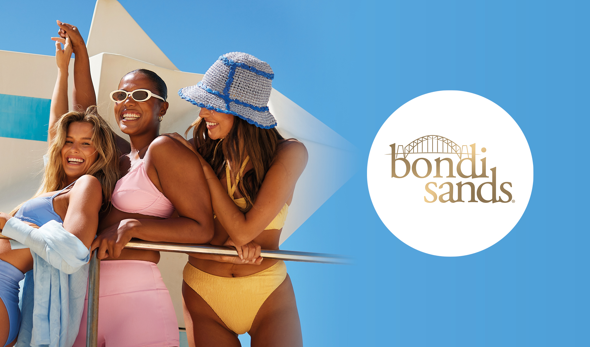 Tre kvinnor klädda i badkläder och stående vid ett räcke utomhus. Den ena kvinnan bär en blå bikini och en ljusblå skjorta, den andra en rosa bikini, och den tredje en gul bikini samt en blå och vit hatt. Bakgrunden visar en klarblå himmel och en vit byggnad. Till höger finns Bondi Sands logotyp i guld på en vit cirkel med en blå bakgrund.