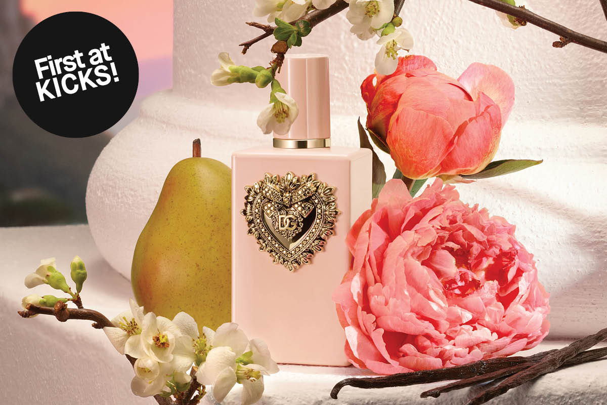 Dolce & Gabbana parfymeflaske i lys rosa med gulldetalj og hjerteformet DG-dekor er omgitt av blomster og frukt på en lys overflate. En svart sirkel viser teksten “First at KICKS!”.