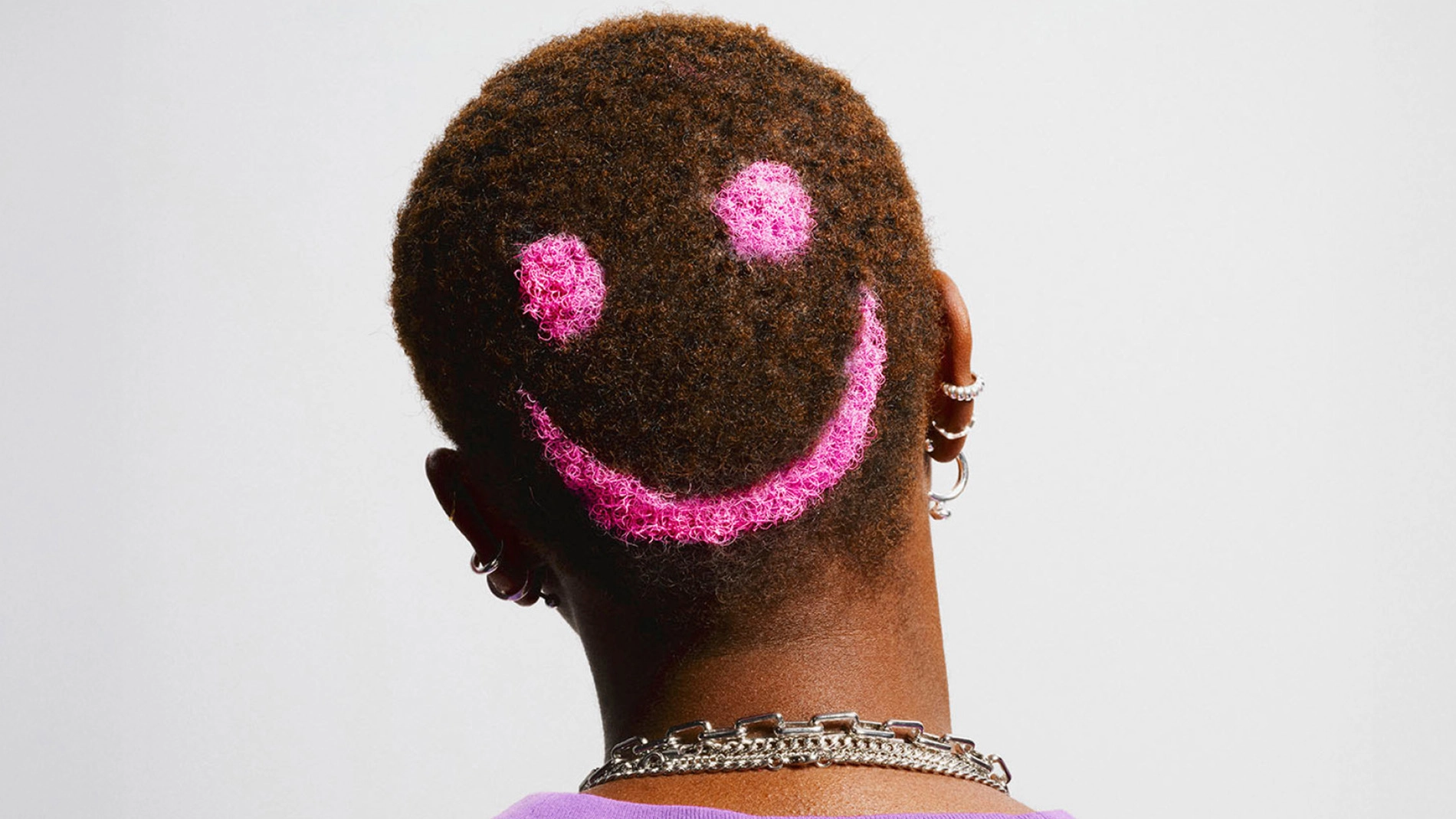 Baksiden av et hode med kort, mørkt hår der en rosa smileyform er laget i håret. Personen har flere øredobber og halskjeder mot en lys bakgrunn.