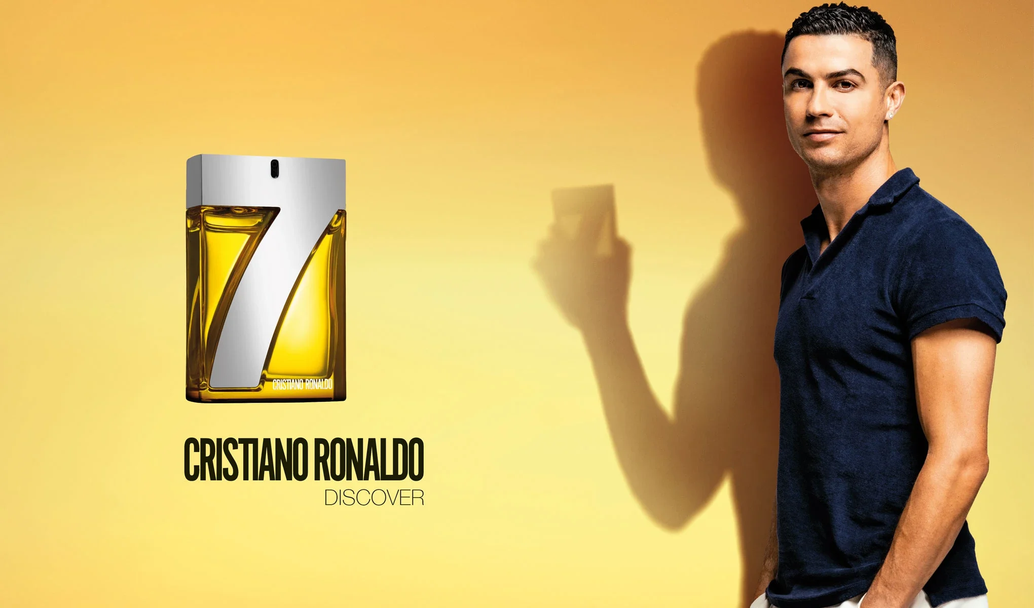 Parfymflaska med texten "Cristiano Ronaldo Discover" och en stor siffra 7 på flaskan. Till höger står en man klädd i en mörkblå pikétröja, mot en gul bakgrund. Mannen håller i en parfymflaska och hans skugga syns tydligt på väggen bakom honom.