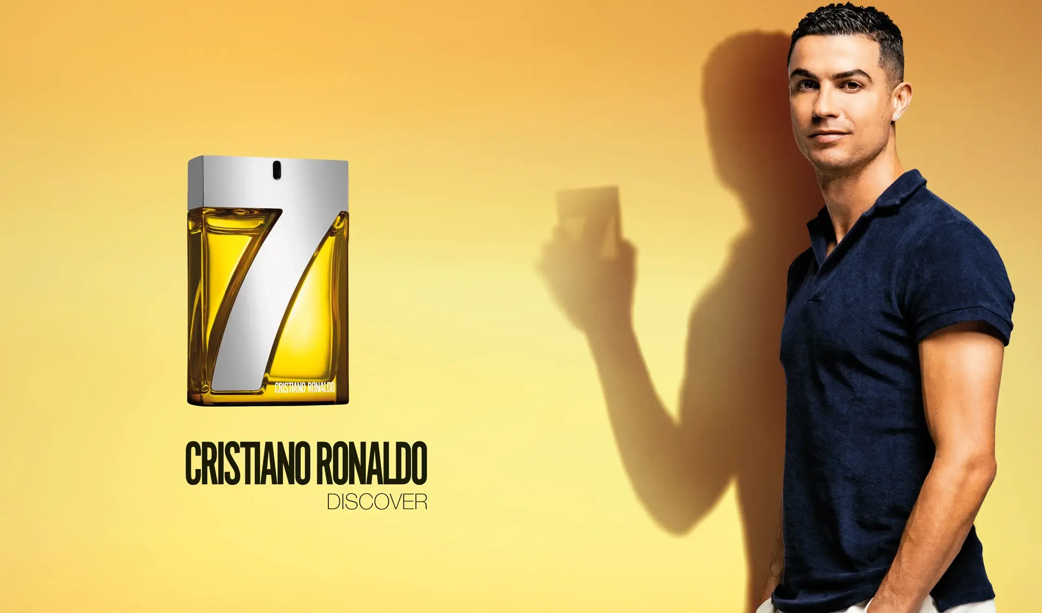 Parfymeflaske med teksten «Cristiano Ronaldo Discover» og et stort nummer 7 på flasken. Til høyre står en mann i mørkeblå poloskjorte mot en gul bakgrunn. Mannen holder en parfymeflaske, og skyggen hans er tydelig synlig på veggen bak ham.