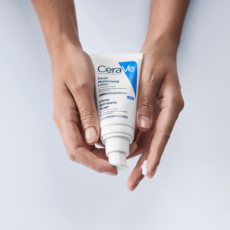 Två händer som håller en flaska CeraVe Facial Moisturising Lotion. En liten mängd lotion har pumpats ut på ena handen. Flaskans etikett är vit med blå och svart text. Bakgrunden är ljusgrå.