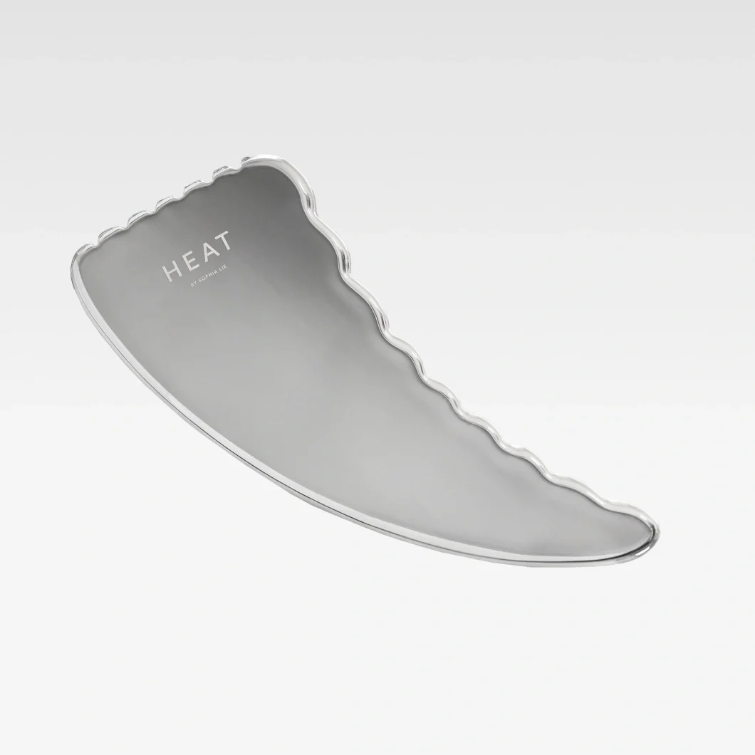 Silverfärgad gua sha från HEAT by Sophia Lie med vågformad kant på ljusgrå bakgrund. Beautyverktyg för ansiktsmassage och hudvård.