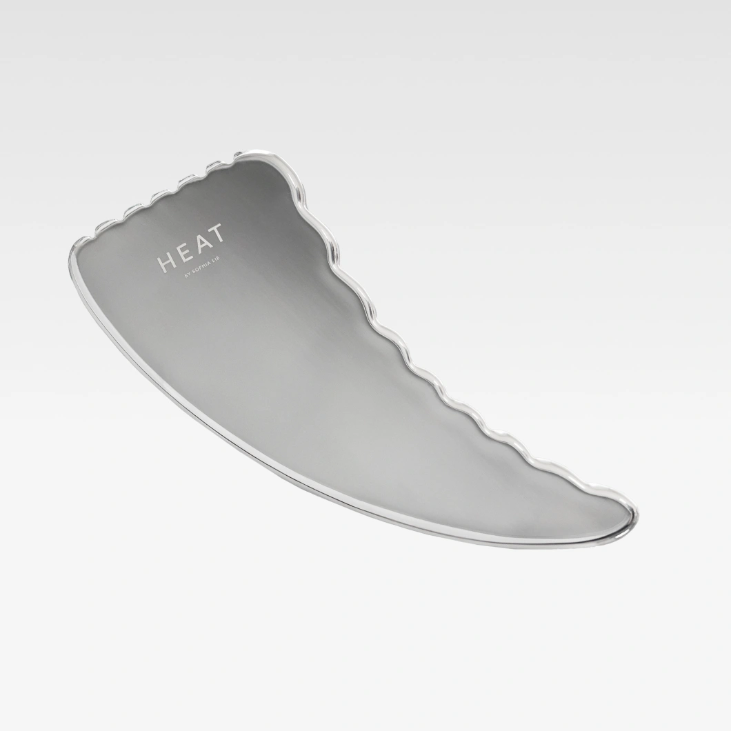 Hopeanvärinen HEAT by Sophia Lie -gua sha aaltoilevalla reunalla vaaleanharmaalla taustalla. Beauty-työkalu kasvohierontaan ja ihonhoitoon.