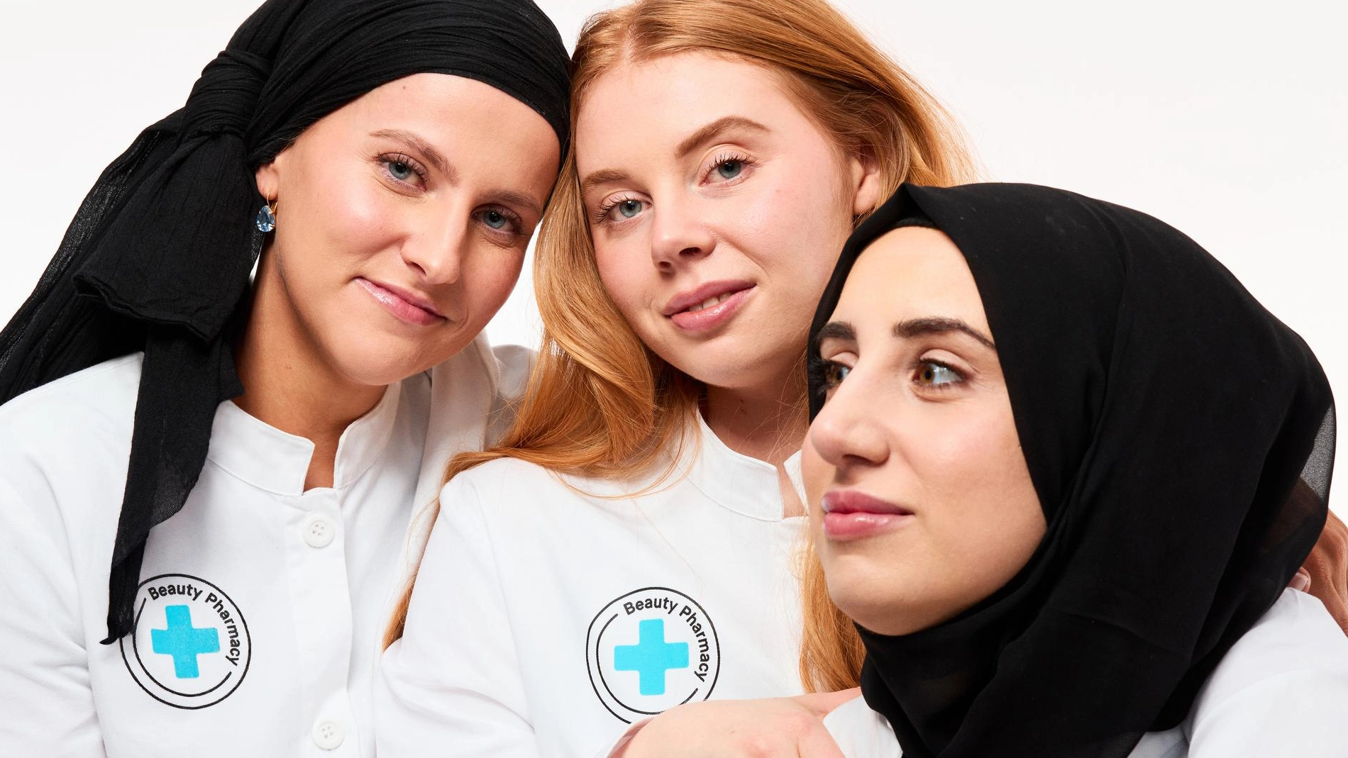 Tre kvinnor i vita arbetskläder från "Beauty Pharmacy" står nära varandra och ler mot kameran, två bär svart sjal.