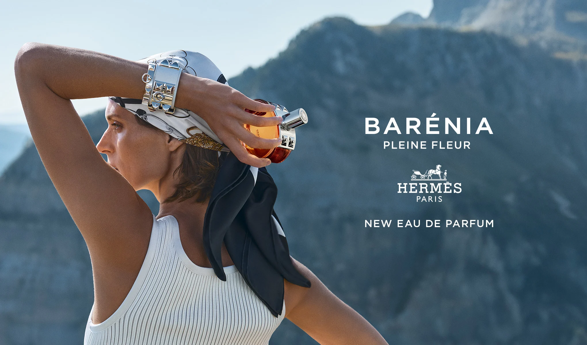Kvinna håller Hermès Barénia Pleine Fleur-parfymflaskan i ett bergslandskap. Text: ”Barénia Pleine Fleur. Hermès Paris. New Eau de Parfum.”