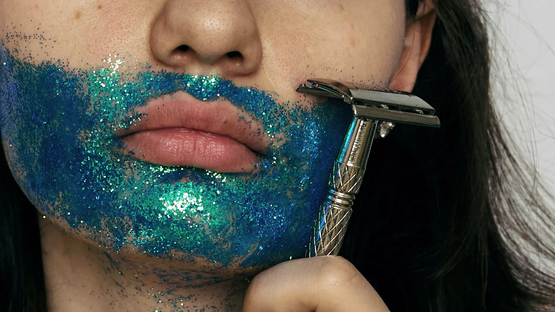 Nærbilde av munn og hake med blågrønt glitter på huden, der en metallbarberhøvel holdes mot haken under barbering mot mørk bakgrunn.