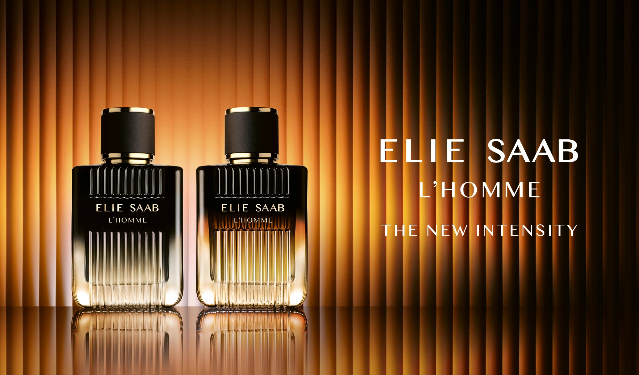Kaksi Elie Saab L’Homme -hajustepulloa musta- ja kullanvärisessä designissa seisoo heijastavalla pinnalla lämpimän raidallisen taustan edessä. Oikealla näkyy teksti “ELIE SAAB L’HOMME” ja “THE NEW INTENSITY”.