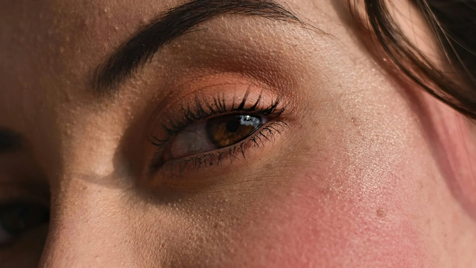 Nærbilde av et øye med rosa øyenskygge, svart mascara og markert mørkt øyenbryn. Rosa rouge er synlig på kinnet.