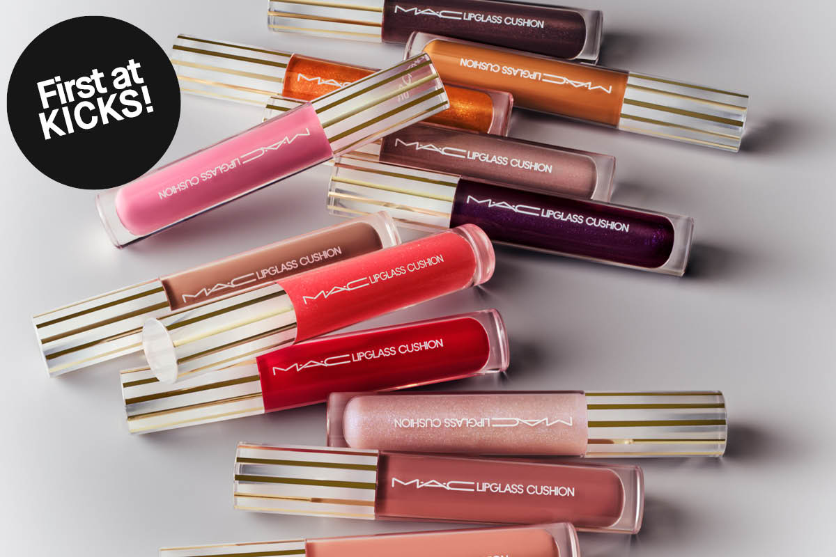 Useita MAC Lipglass Cushion -huulikiiltoja eri sävyissä on aseteltu vaalealle taustalle, kultaraitaisilla korkilla. Tuotteissa lukee “MAC Lipglass Cushion” ja mustassa ympyrässä “First at KICKS!”.
