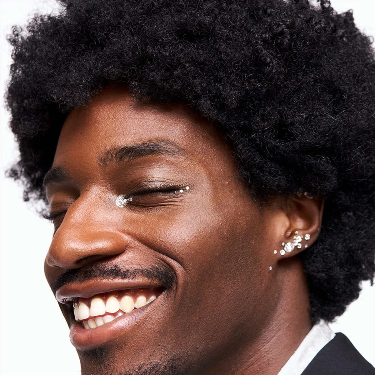 Kuva, joka näyttää henkilön, jolla on mustat kiharat hiukset ja useita korvakoruja korvassa. Henkilöllä on yllään musta takki ja valkoinen paita. Tausta on valkoinen.