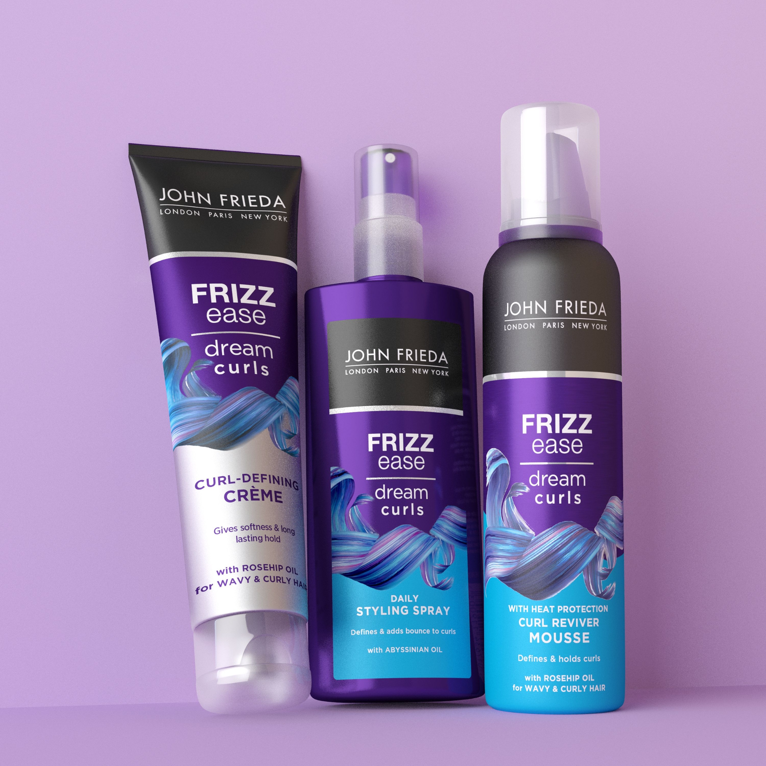 Kolme John Frieda Frizz Ease Dream Curls -tuotetta vaaleanvioletin taustan edessä. Kuvassa on kiharoita muotoileva voide, muotoilusuihke ja kiharoita elvyttävä vaahto.